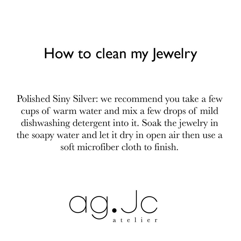 May include: Instructions on how to clean jewelry made of polished silver. The text recommends soaking the jewelry in soapy water and then drying it in open air. The text also recommends using a soft microfiber cloth to finish cleaning the jewelry. The text is followed by the logo "ag.Jc atelier".