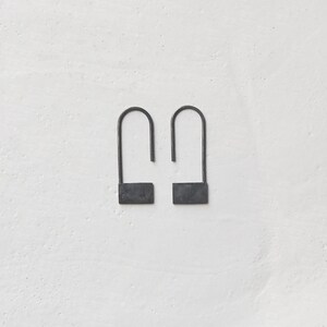Pode incluir: Um par de brincos de metal preto com um design retangular simples. Os brincos são feitos de um fio fino que é dobrado em um laço na parte superior e depois forma um retângulo na parte inferior.