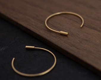 May include: Two gold hoop earrings with a cylindrical closure. The earrings are on a wooden surface and a black surface.