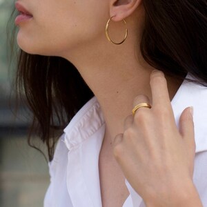 May include: A woman wearing a white shirt with gold hoop earrings and a gold ring on her finger.