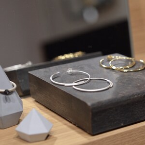 May include: Two silver hoop earrings on a gray display stand. The earrings are stacked on top of each other. The stand is made of a dark gray material and has a rectangular shape.