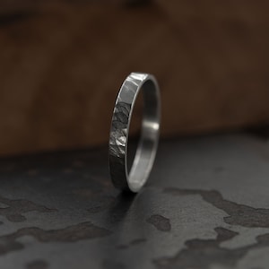 May include: A silver band ring with a hammered texture. The ring is on a dark grey surface.
