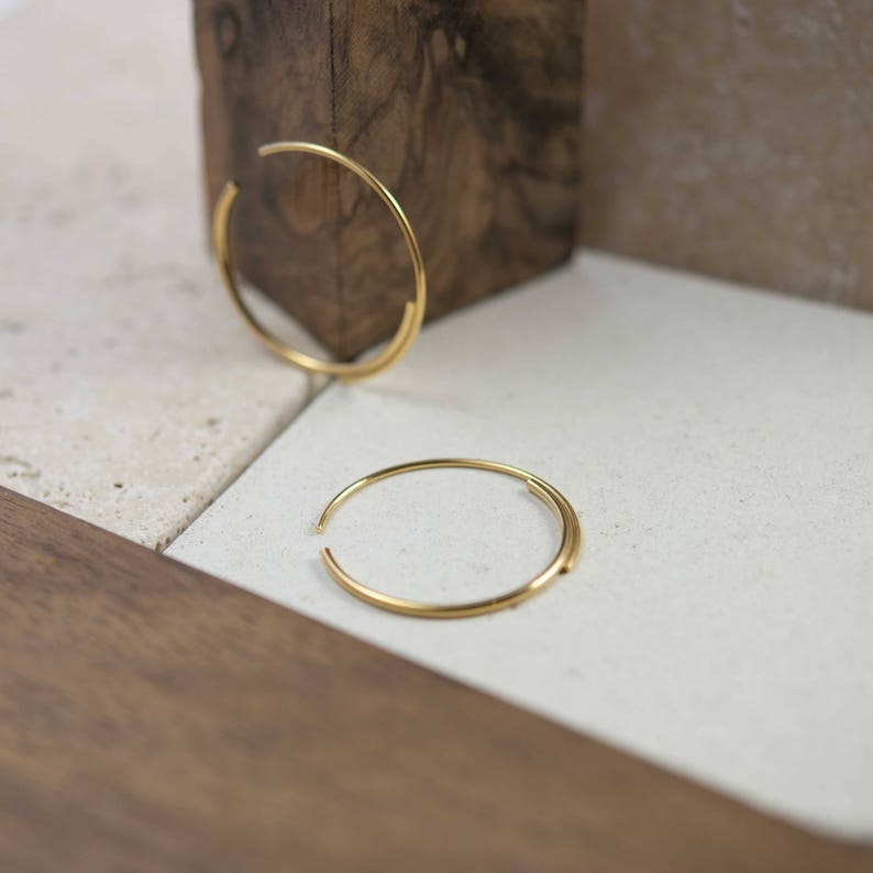 May include: Two gold hoop earrings, one larger than the other, are displayed on a light brown surface. The earrings are made of a thin, round wire.