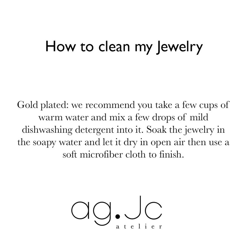 May include: Instructions on how to clean gold-plated jewelry. The text recommends soaking the jewelry in warm water with a few drops of mild dishwashing detergent, then drying it in open air and polishing with a microfiber cloth. The text is followed by the brand name "ag.Jc atelier".