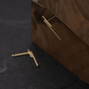 May include: A pair of gold bar earrings. The earrings are simple and minimalist, with a straight bar design. The earrings are shown on a dark wooden surface.