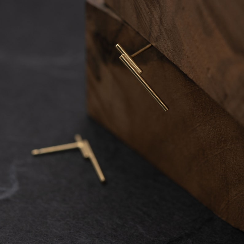 May include: A pair of gold bar earrings. The earrings are simple and minimalist, with a straight bar design. The earrings are shown on a dark wooden surface.