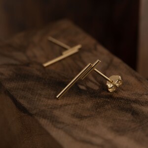 May include: A pair of gold bar stud earrings. The earrings are made of two thin bars that intersect at a right angle. The earrings are on a wooden surface.