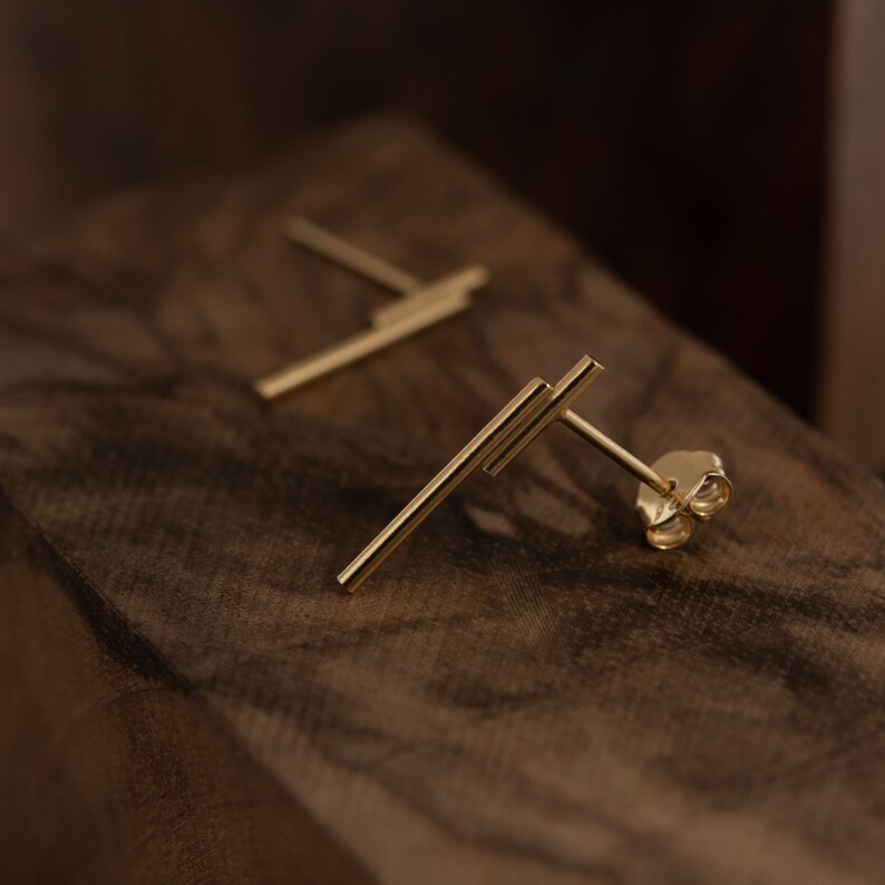 May include: A pair of gold bar stud earrings. The earrings are made of two thin bars that intersect at a right angle. The earrings are on a wooden surface.