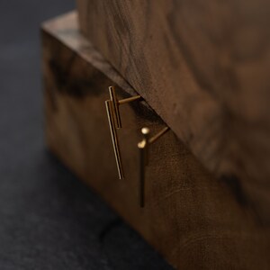 May include: A pair of gold bar earrings with a minimalist design. The earrings are shown on a wooden surface.