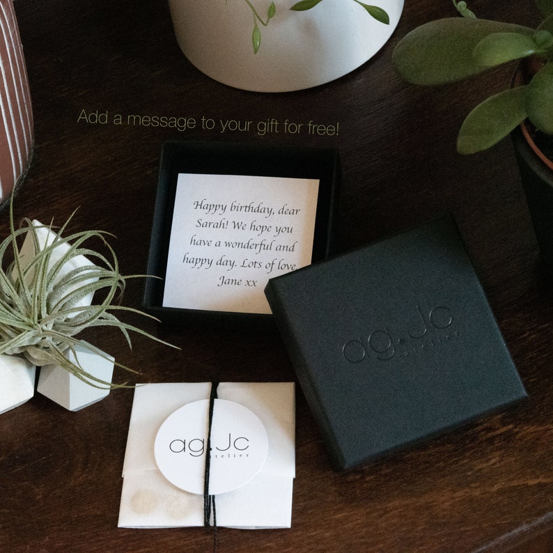 May include: A black gift box with a white card inside. The card has a handwritten message that reads "Happy birthday, dear Sarah! We hope you have a wonderful and happy day. Lots of love Jane xx". The box has a black label with the text "ag Jc" in white.