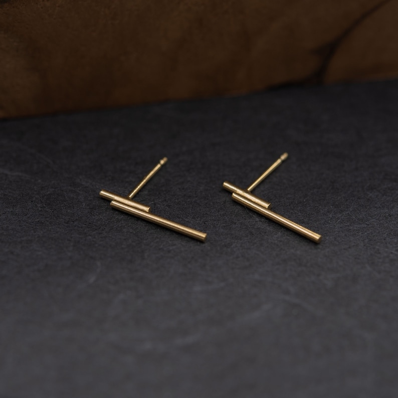 May include: A pair of gold bar earrings with a minimalist design. The earrings are made of two thin bars of gold that are stacked on top of each other.