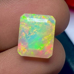 May include: A rectangular opal gemstone displaying a vibrant play of colour, including green, yellow, and red hues. The gemstone is held between fingers, showcasing its polished surface and internal iridescence. The opal is a gemstone.