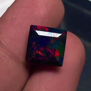 May include: A square-cut black opal gemstone with vibrant internal colours of red, green, and blue. The gemstone is held between fingers, showcasing its polished surface and play of colour. The opal is a gemstone.