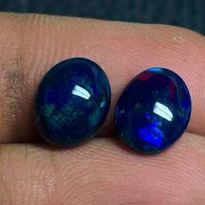Puede incluir: Dos piedras preciosas ovaladas pulidas, de color azul profundo con destellos iridiscentes verdes y azules. Las piedras se sostienen entre los dedos, mostrando sus superficies reflectantes y su belleza natural. Probablemente ópalos.