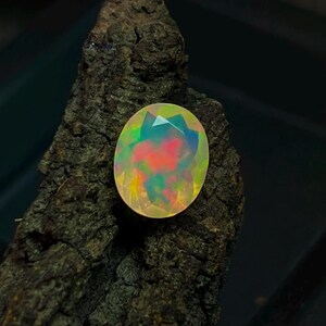 May include: An oval opal gemstone with a faceted cut, showcasing a vibrant play of colour including red, green, and yellow hues. The gemstone is set against a dark, textured, wooden surface, creating a striking contrast.