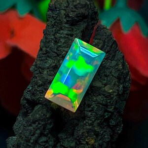 Puede incluir: Una gema rectangular con colores vibrantes de verde, azul y amarillo. La gema se presenta sobre un fondo oscuro y texturizado, resaltando su brillo. El corte y pulido de la gema crean una cautivadora exhibición de luz y color.