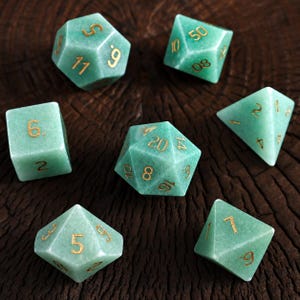 May include: A set of seven polyhedral dice crafted from light green stone with a matte finish. Each die features engraved numbers filled with gold-colored ink, including a twenty-sided die, a twelve-sided die, two ten-sided dice, an eight-sided die, a six-sided die, and a four-sided die.