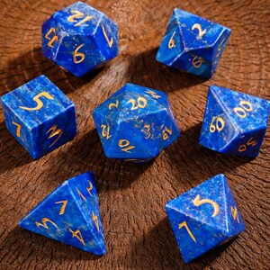 以下が含まれることがあります： 金色の数字が刻まれた、青い宝石で作られた7つのサイコロのセット。d4、d6、d8、d10、d12、d20、およびパーセンテージサイコロが含まれています。サイコロは木製の表面に置かれ、その多面的な形状と複雑なディテールが示されています。