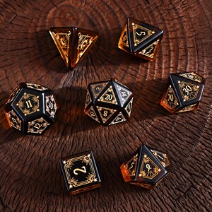 Puede incluir: Un juego de siete dados poliédricos con un diseño degradado de ámbar translúcido y negro opaco. Cada dado está adornado con intrincados grabados dorados de inspiración celestial, que incluyen patrones de rayos de sol y bordes geométricos alrededor de los números.