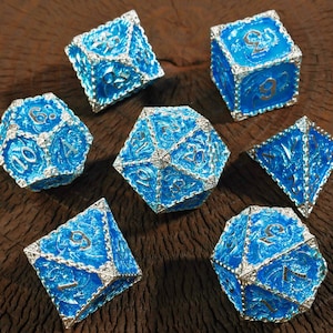 May include: Set of seven polyhedral dice in various shapes, including a d4, d6, d8, d10, d12, d20, and a percentile die. The dice are a vibrant blue with silver-colored trim and gold-colored numbers. The dice are displayed on a dark brown wooden surface.
