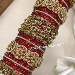 May include: A stack of ornate red and gold bangles. The bangles feature intricate floral and geometric designs with embedded gemstones and pearls. The text "Adventure shop gifts" is visible.