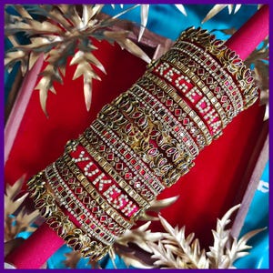 May include: A collection of ornate red and gold bangles, adorned with intricate patterns and sparkling embellishments. The bangles are arranged on a red cylindrical base, with gold floral accents. The background features gold leaf-like decorations and a vibrant blue surface.
