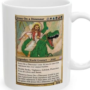 May include: White ceramic mug with a humorous illustration of Jesus riding a green dinosaur. The mug features the text "Jesus On a Dinosaur" and game-related text. The mug has a curved handle.