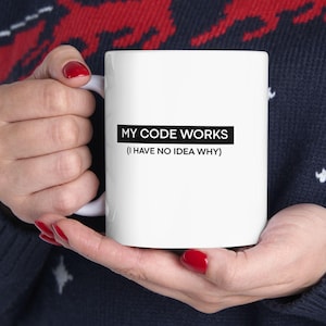 May include: White ceramic mug with the text "MY CODE WORKS (I HAVE NO IDEA WHY)" in black. The mug is held by a person wearing a dark blue sweater with a red and white reindeer pattern.