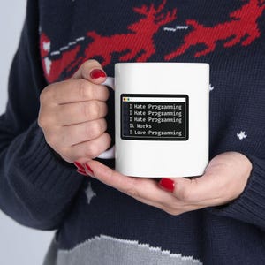 May include: White ceramic mug with the text "I Hate Programming, I Love Programming" displayed in a black computer terminal style. The mug is held by a person wearing a dark blue sweater with a reindeer pattern.