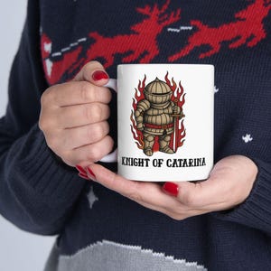 May include: White ceramic mug featuring a cartoon knight in full armor, holding a sword, surrounded by flames. The text "KNIGHT OF CATARINA" is printed below the illustration. The mug is held by a person wearing a dark blue sweater.