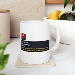 Puede incluir: Taza de cerámica blanca con un diseño gráfico rectangular negro. El diseño incluye el texto "Will to Live", "Unlimited range" e "Instant". La taza también presenta un icono de calavera roja y texto adicional sobre la curación.