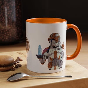 Op de afbeelding: Witte keramische mok met een oranje binnenkant en handvat. De mok heeft een gedetailleerde illustratie van een personage in een wit en oranje uniform dat een blauw object vasthoudt. Aan de linkerkant van de mok staat een bruin logo.