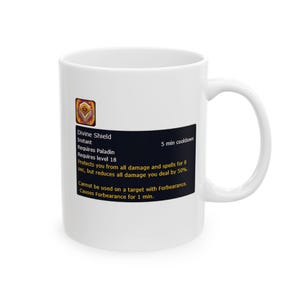 Puede incluir: Taza de cerámica blanca con el texto "Divine Shield" y detalles del juego. La taza tiene un rectángulo negro con texto blanco y un icono dorado y naranja. La taza tiene un asa curva.