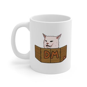Könnte beinhalten: Weiße Keramik-Tasse mit einem Meme einer Katze hinter einem Holzschirm mit den Buchstaben "DM" in Orange. Die Katze hat einen selbstgefälligen Gesichtsausdruck. Die Tasse hat einen gebogenen Griff.