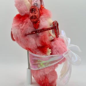 May include: A whimsical pink and red plush creature with glittery accents. It wears red glitter glasses and has a white bow. The creature is on a white stand, showcasing its unique design and playful aesthetic.