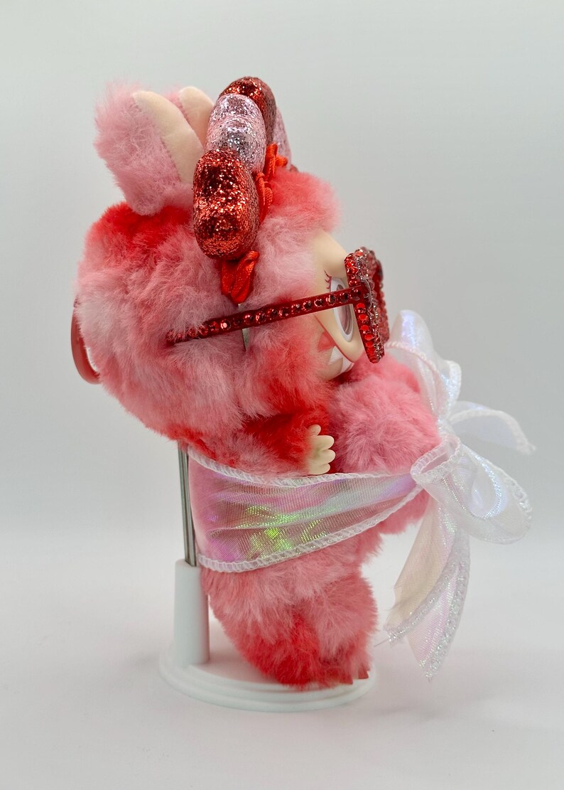 May include: A whimsical pink and red plush creature with glittery accents. It wears red glitter glasses and has a white bow. The creature is on a white stand, showcasing its unique design and playful aesthetic.