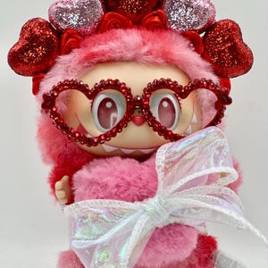 May include: A pink plush figure with heart-shaped glasses and a heart-adorned headpiece. The figure has a large white bow and a tag that reads "THEM". The figure is a Valentine's Day themed collectible.