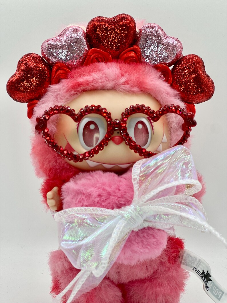 May include: A pink plush figure with heart-shaped glasses and a heart-adorned headpiece. The figure has a large white bow and a tag that reads "THEM". The figure is a Valentine's Day themed collectible.