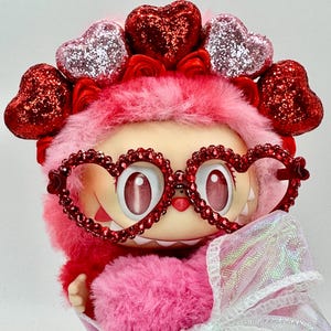 May include: A plush, pink creature with heart-shaped glasses and a matching headband. The glasses and headband are adorned with red glitter hearts. A white, iridescent bow is tied around the creature's neck. The creature has large, expressive eyes.