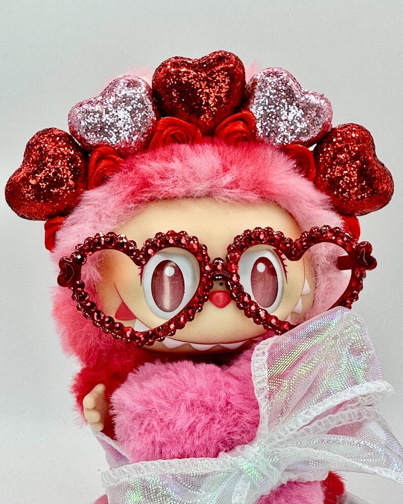May include: A plush, pink creature with heart-shaped glasses and a matching headband. The glasses and headband are adorned with red glitter hearts. A white, iridescent bow is tied around the creature's neck. The creature has large, expressive eyes.