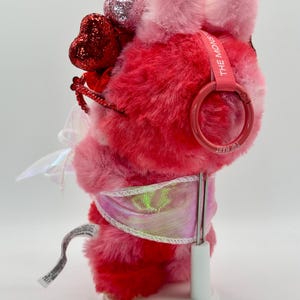 May include: A plush, heart-themed toy in shades of pink and red, adorned with glittery heart accents. It features a pink strap with the text "THE MONS" and is displayed on a white stand. The toy has a fluffy texture and a whimsical design.