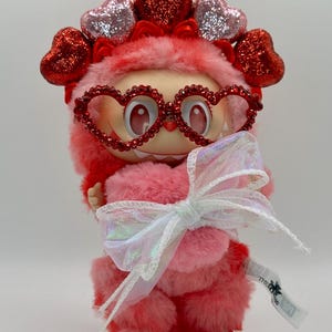 May include: A plush pink figure with a heart-shaped headpiece and heart-shaped glasses. The figure has a fluffy body and holds a white iridescent bow. The figure is on a white base.