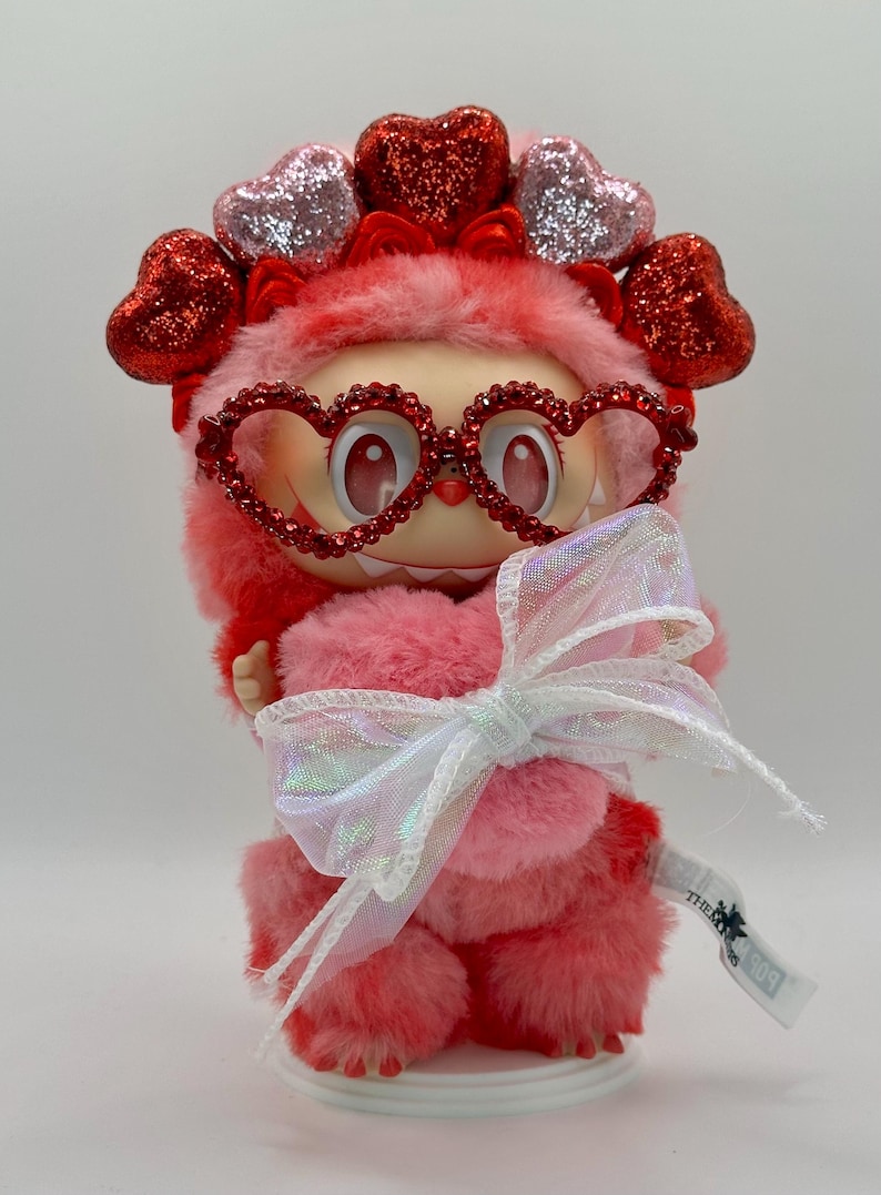 May include: A plush pink figure with a heart-shaped headpiece and heart-shaped glasses. The figure has a fluffy body and holds a white iridescent bow. The figure is on a white base.