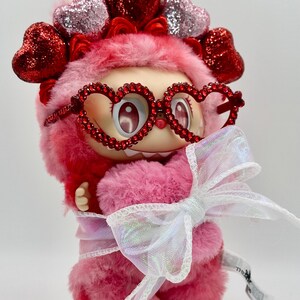 May include: A pink plush figure with heart-shaped glasses and a matching headband. The figure is adorned with a large white bow and has a fluffy texture. The overall design is whimsical and cute, with a focus on Valentine's Day.