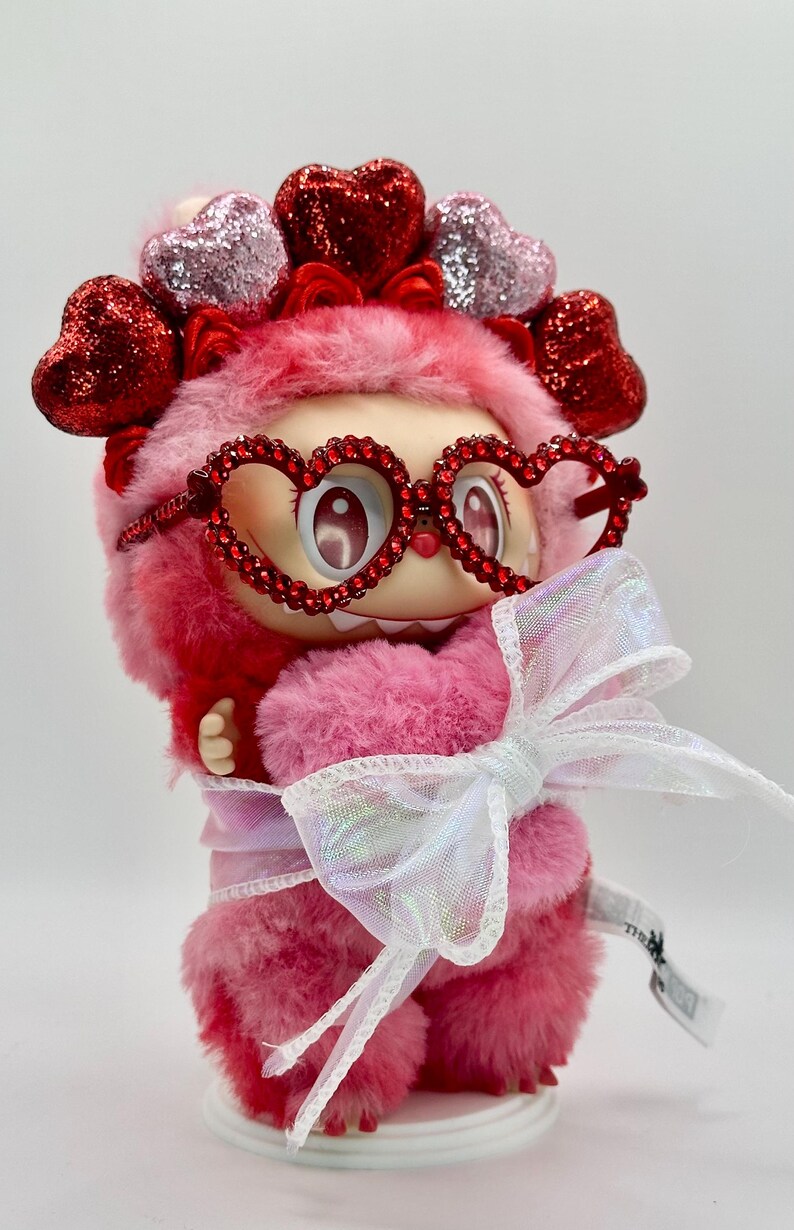 May include: A pink plush figure with heart-shaped glasses and a matching headband. The figure is adorned with a large white bow and has a fluffy texture. The overall design is whimsical and cute, with a focus on Valentine's Day.