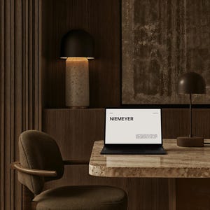 Op de afbeelding: Een moderne kantoorruimte met een laptop met de tekst "NIEMEYER", een bureaulamp en een decoratieve lamp. Het bureau heeft een gespikkeld oppervlak en een bruine stoel staat op de voorgrond. De achtergrond is donker hout.