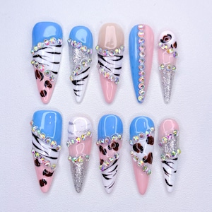 May include: A set of ten artificial nails in various designs. The nails feature a mix of colors, including blue, pink, and nude, with accents of glitter, rhinestones, and animal print patterns. Each nail has a unique design.