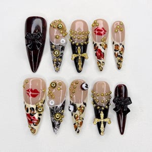 May include: A collection of ten long, almond-shaped artificial nails. Designs include a dark burgundy nail with a black bow, nails with gold embellishments, a cross, and a lip design. Some nails feature leopard print and pearls.