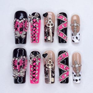 May include: A collection of ten artificial nails with diverse designs. The nails showcase black, pink, and nude bases, embellished with silver bows, rhinestones, key charms, and cross details. Some nails have lace patterns and a cow print.