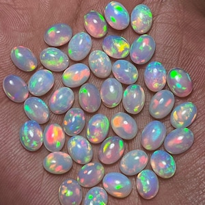 Puede incluir: Una colección de ópalos ovalados, cada uno con una base blanca lechosa y destellos iridiscentes de verde, rosa y amarillo. Las piedras preciosas están dispuestas muy juntas, mostrando su juego de color y brillo únicos. Estos ópalos son ideales para la fabricación de joyas.
